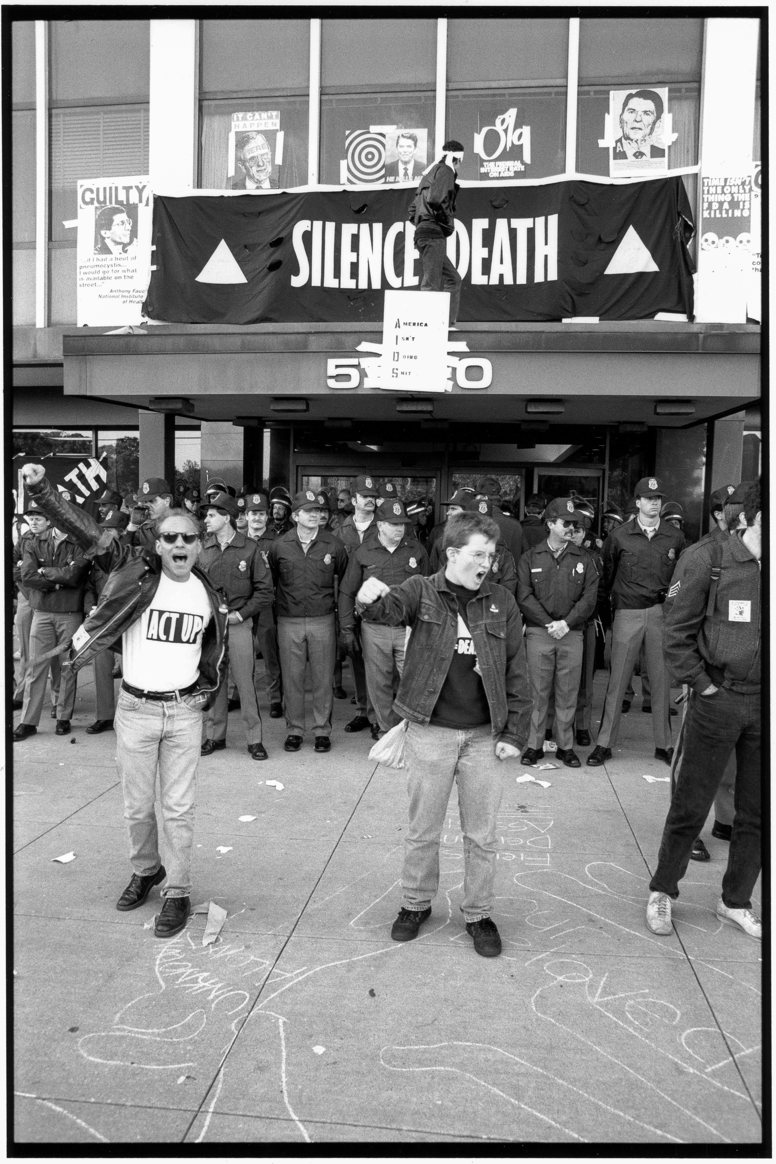 ACT UP protesters close the Federal Drug Administration building to demand the release of experimental medication for those living with HIV/AIDS. Their slogans read: “Silence=Death,” and “AIDS: America Isn't Doing Shit.” The demonstration was held outside the FDA headquarters in Rockville, Maryland, on October 11, 1988.