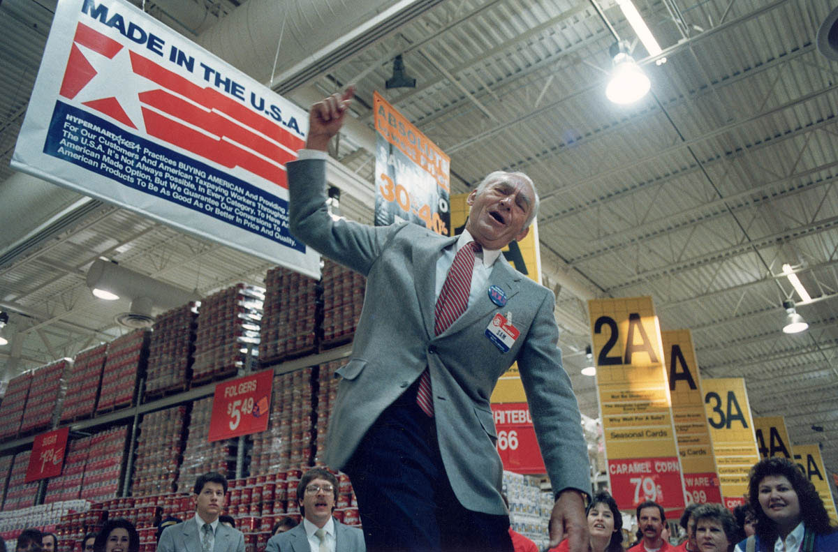 Using the crowd-pleasing tactics of a high school pep rally, Sam Walton exhorts employees at the opening of a new Wal-Mart “Hypermart.”